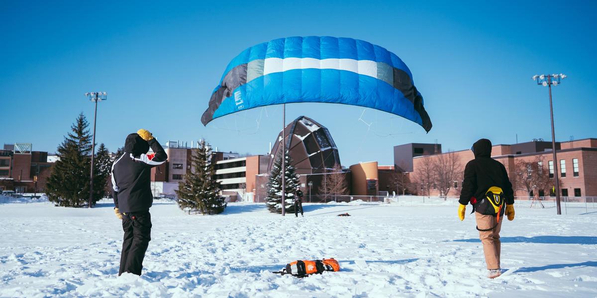 Students learn snowkiting on the University of Minnesota Duluth campus. A large blue foil kite rises against a blue sky with Weber Music Hall in the background.