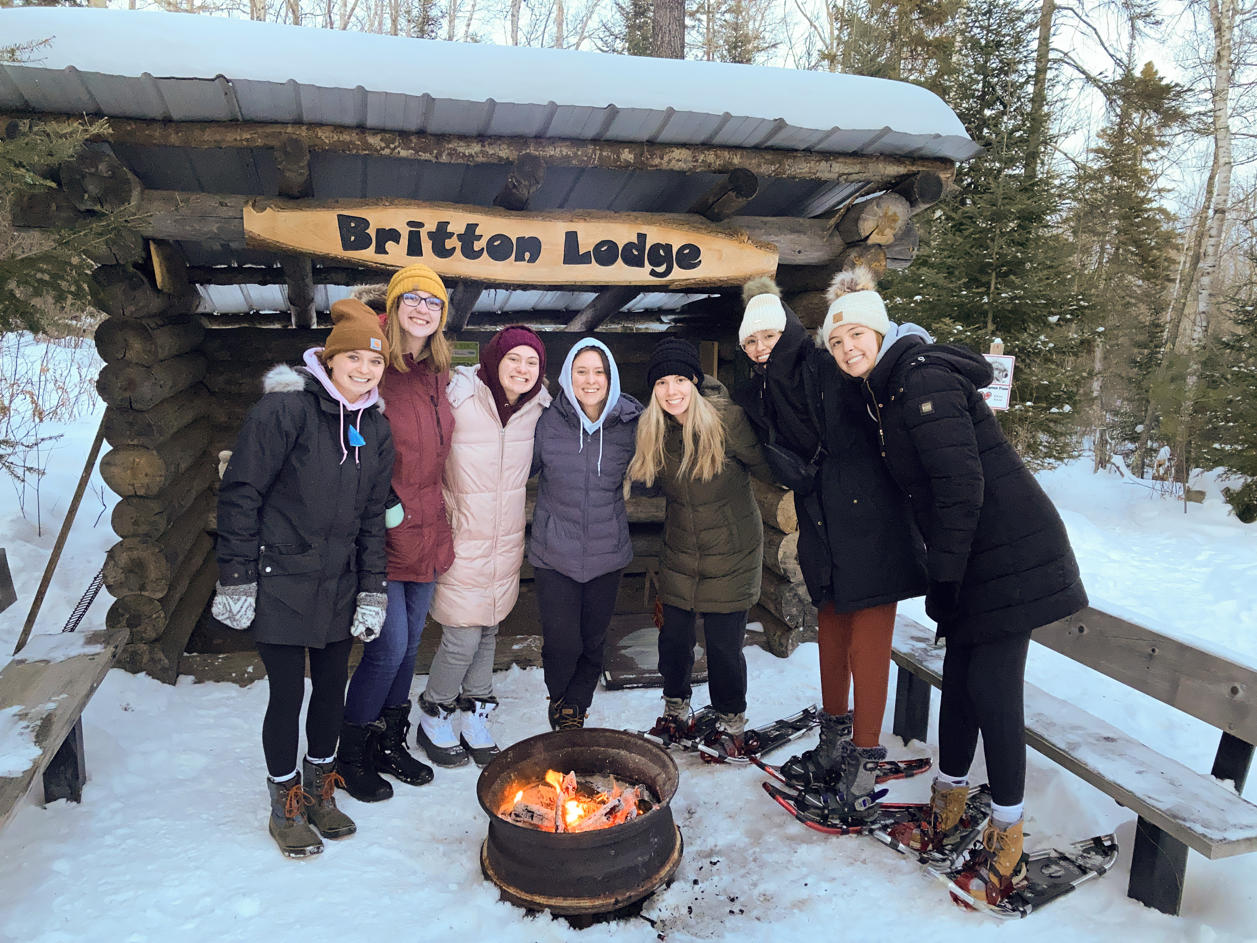 A group of college students in front of a campfire, with a sign in the background reading "Britton Lodge."