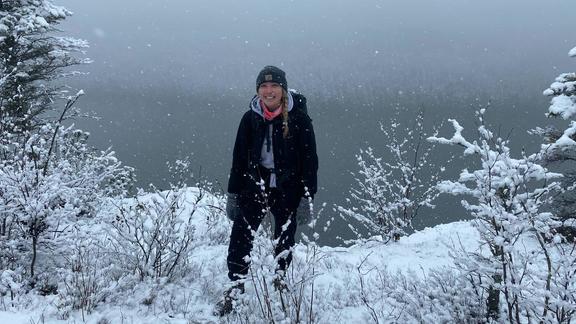 A person in winter clothing standing in the snow near a lake.