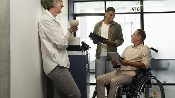 Three people, two standing and one sitting in a wheelchair, having a conversation in an office setting