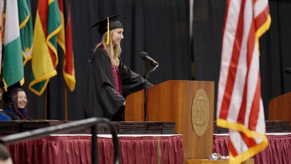 Amanda Fowler speaking at the podium on the commencement stage, wearing a cap and gown, with many flags and university officials behind her