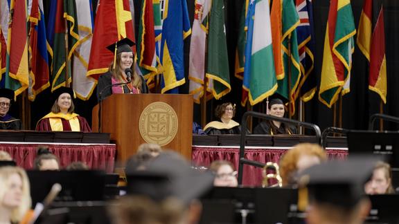 Gavi Gunther speaking on the Commencement stage wearing a cap and gown with many flags and university officials behind her