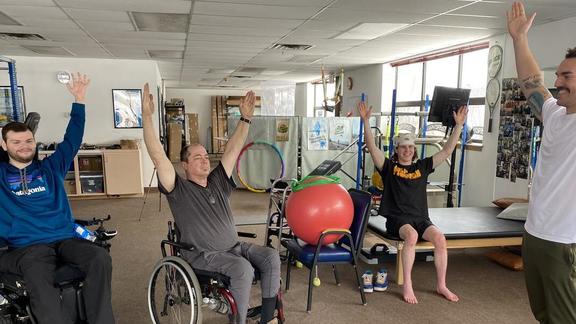 Four people doing a hand raise exercise in an exercise room, one standing, one sitting, and two in a wheel chair.