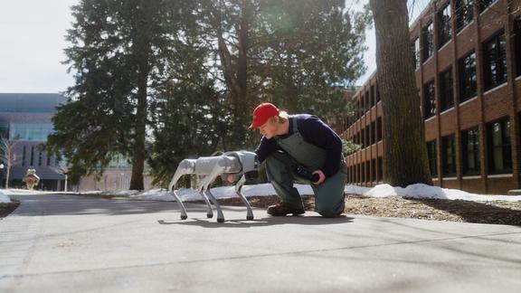 Student Braden Kowalski kneels next to a robotic dog on a sidewalk on the University of Minnesota Duluth campus.