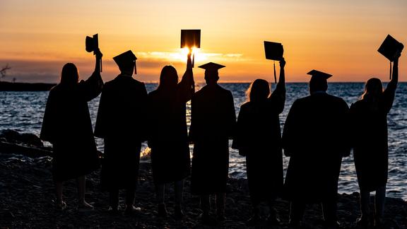 A row of college graduates raise their commencement caps. They&#039;re silhouetted against a sunrise and backdrop of Lake Superior
