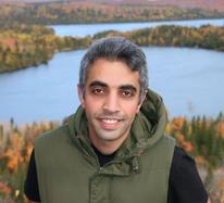 A photo of Siavash Sattar with a green vest in front of lakes and autumn trees.