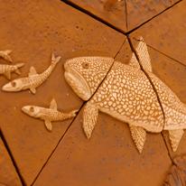 A detailed image of a ceramic artwork shows a lake trout chasing smelt.