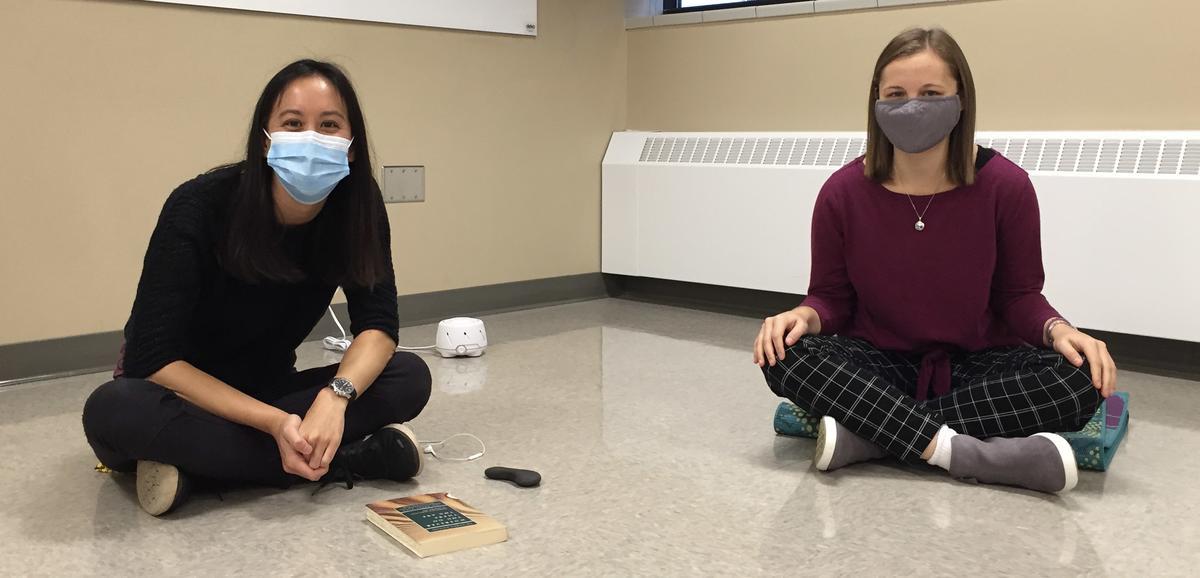 Two students wearing masks, sitting on the floor with legs crossed.