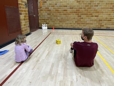 An adult and a student sitting on the floor of a gym, near some bowling pins.