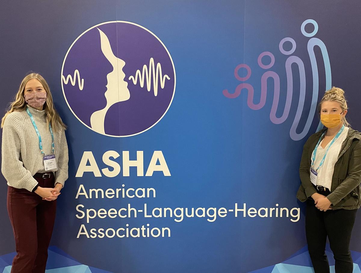 Two woman standing next to a background sign reading "ASHA American Speech-Language-Hearing Association."