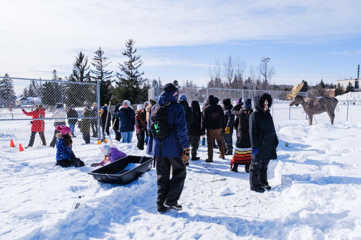 A group of people standing in the snow, some with sleds. There is a fake moose in the background.