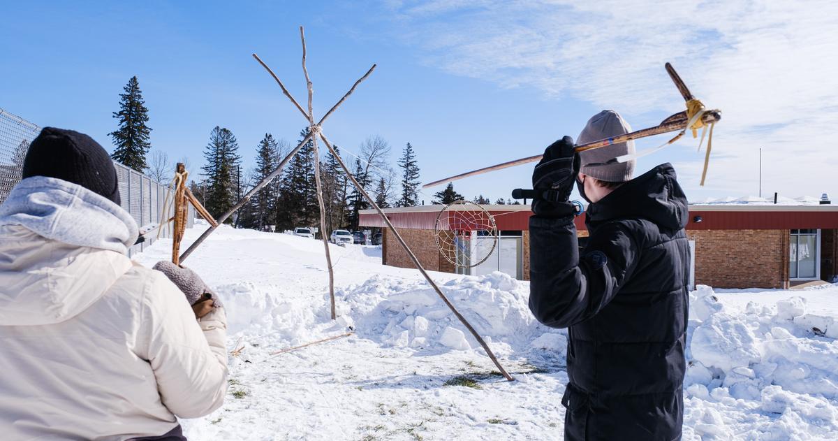 Two people standing in the snow. One is getting ready to throw a spear into a hoop that is hanging from a tripod of sticks.
