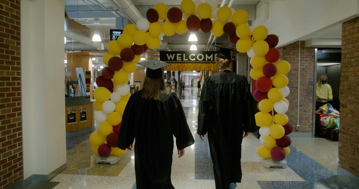 Two students in graduation regalia walk under balloon arch in Kirby