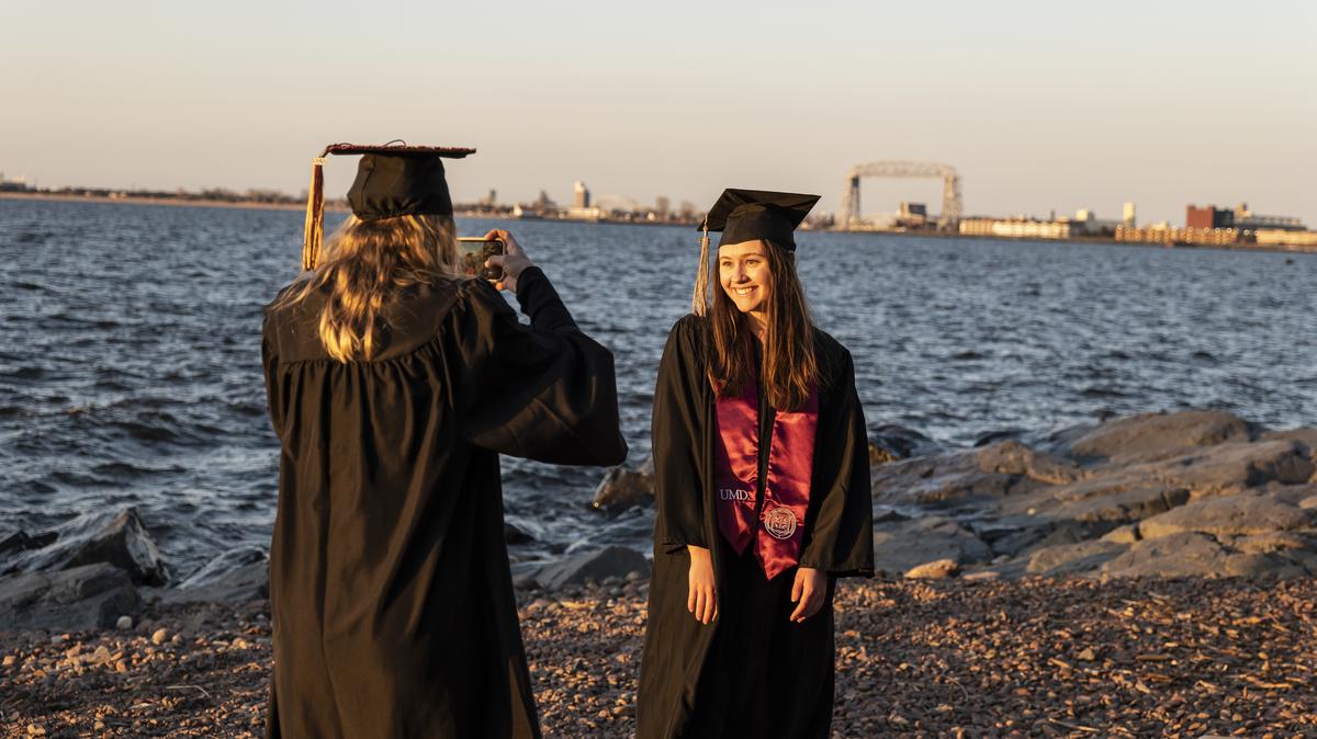 Two students on the beach in graduation regalia taking a picture