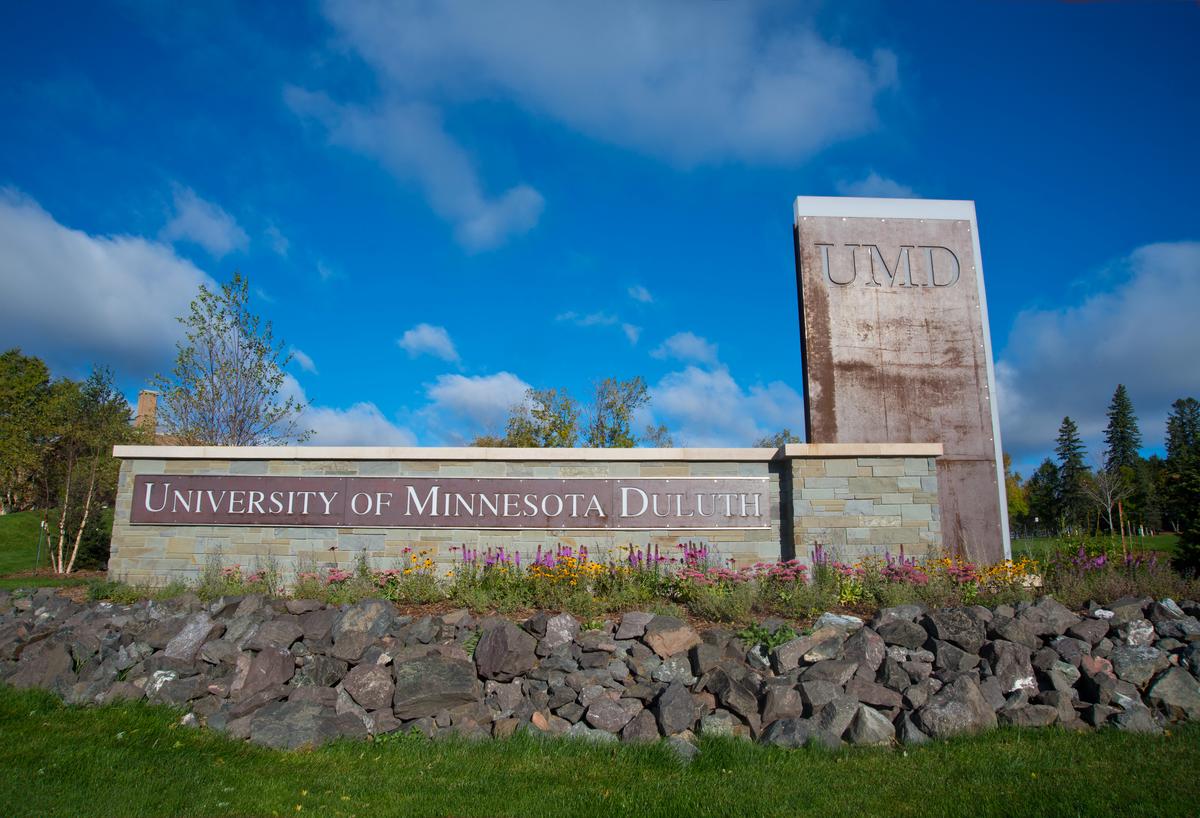 The University of Minnesota Duluth monument sign at Woodland and College under a deep blue sky.
