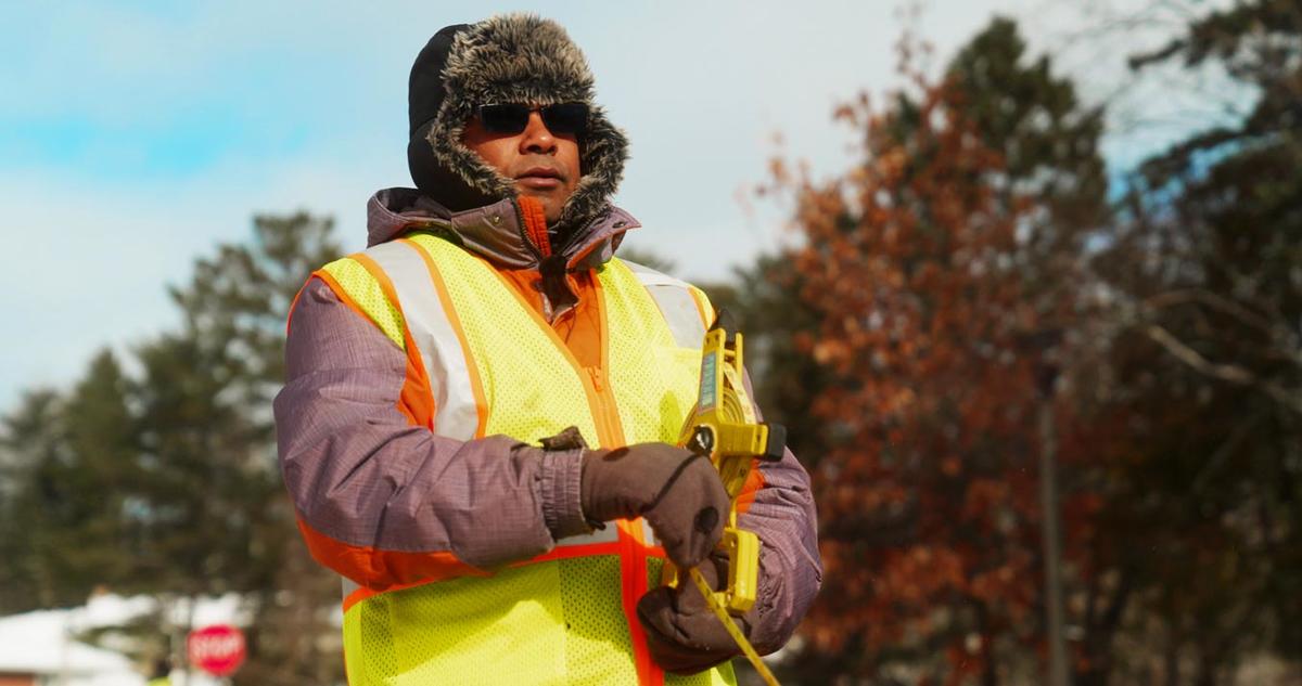 Manik Barman, Ph.D, conducts pothole and road infrastructure field research near UMD while wearing a furry hat, reflective vest and measuring tape.