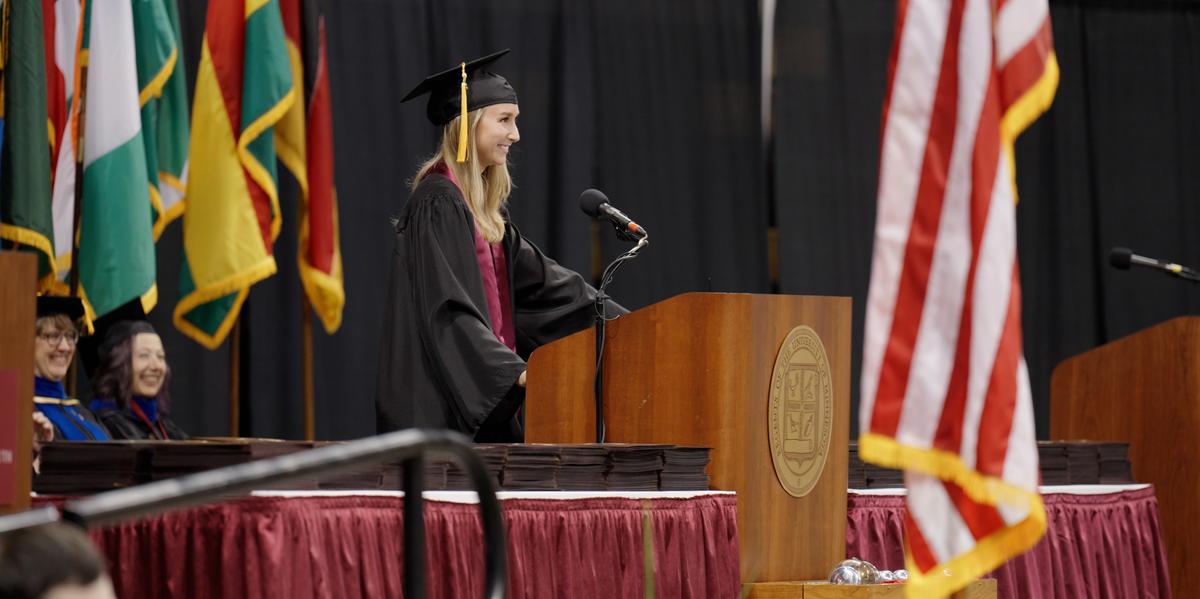 Amanda Fowler speaking at the podium on the commencement stage, wearing a cap and gown, with many flags and university officials behind her