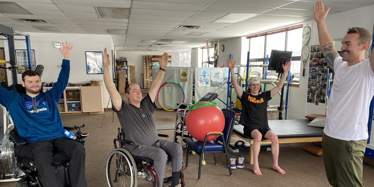 Four people doing a hand raise exercise in an exercise room, one standing, one sitting, and two in a wheel chair.