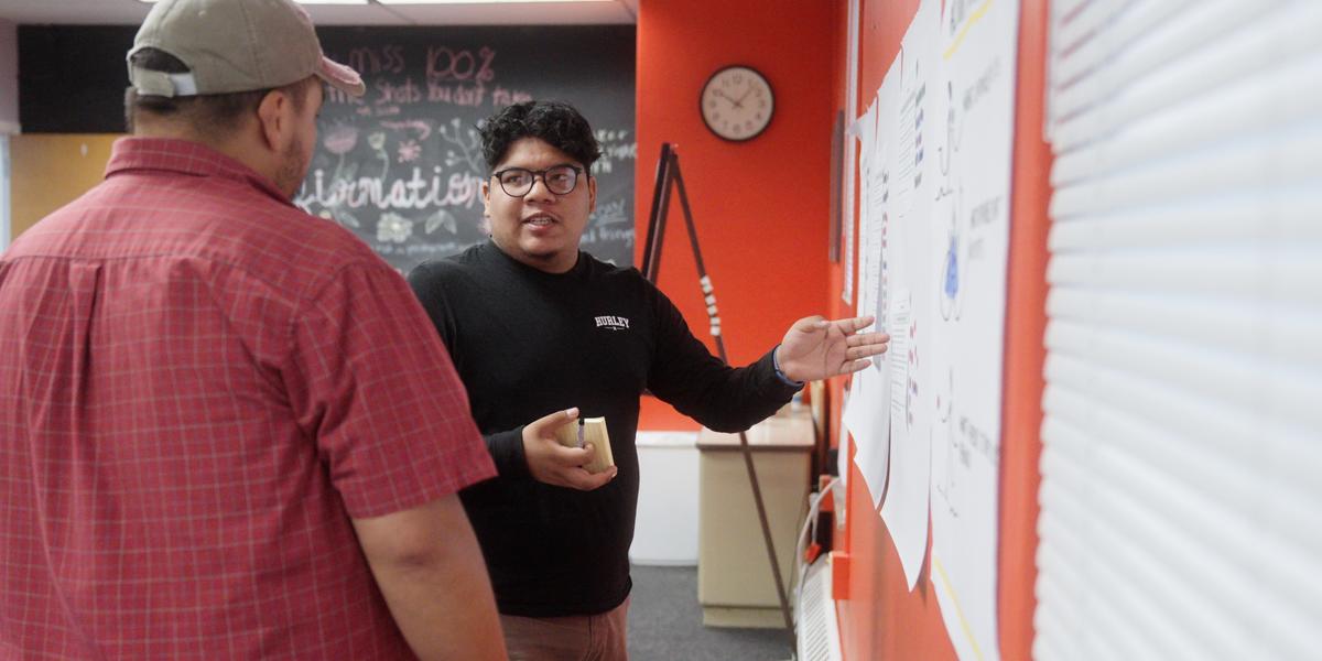Johan Lopez-Ortega stands and gestures towards an idea board while chatting with a colleague