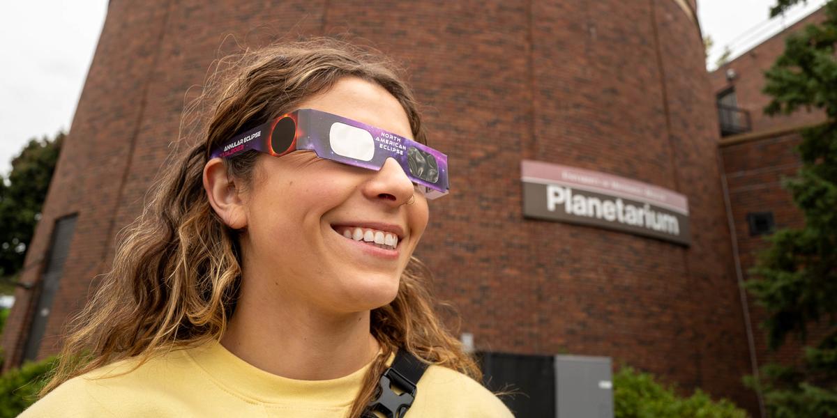 A woman wearing eclipse sunglasses smiles in front of the UMD Planetarium