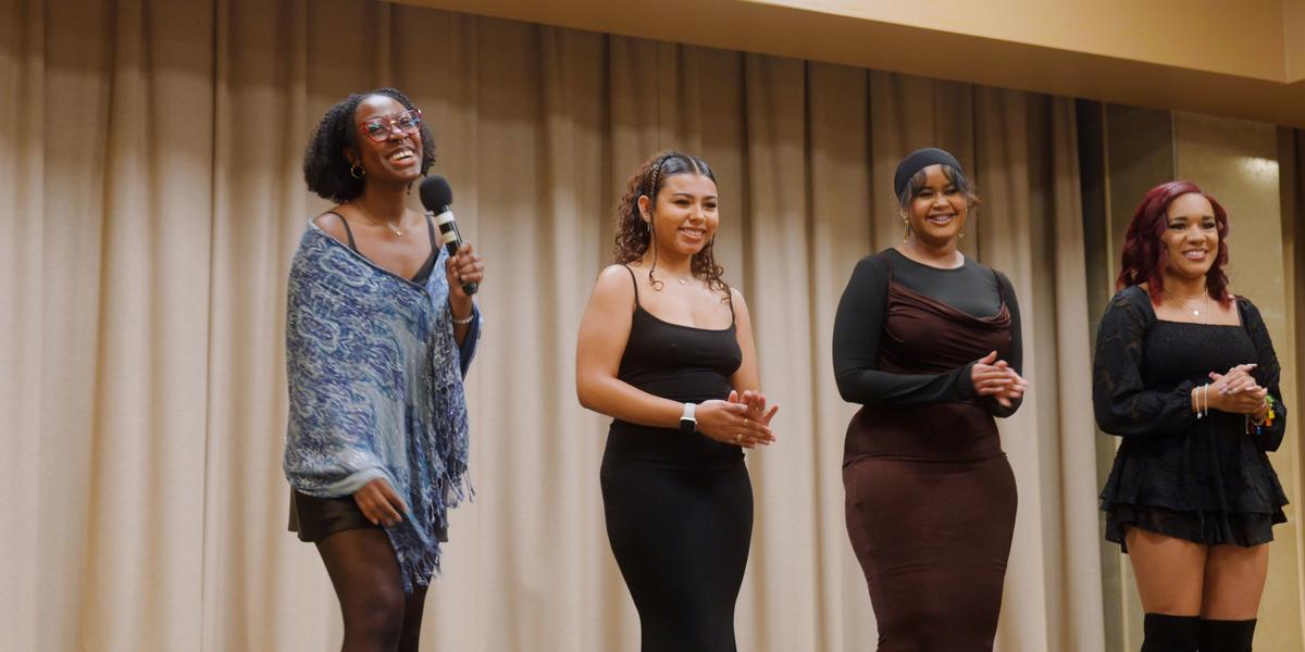 Elizabeth Manly-Spain stands on stage, smiling and holding a microphone, with fellow UMD Black Student Association members at the annual Soul Food Dinner.