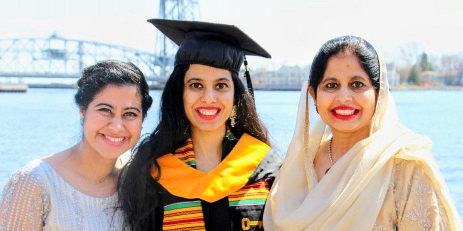 Madiha Shakil Mirza and her sister and mother smile at the camera in front of a sunny Duluth Aerial Lift Bridge. Madiha is wearing a cap and gown, while the other two are wearing white dresses.