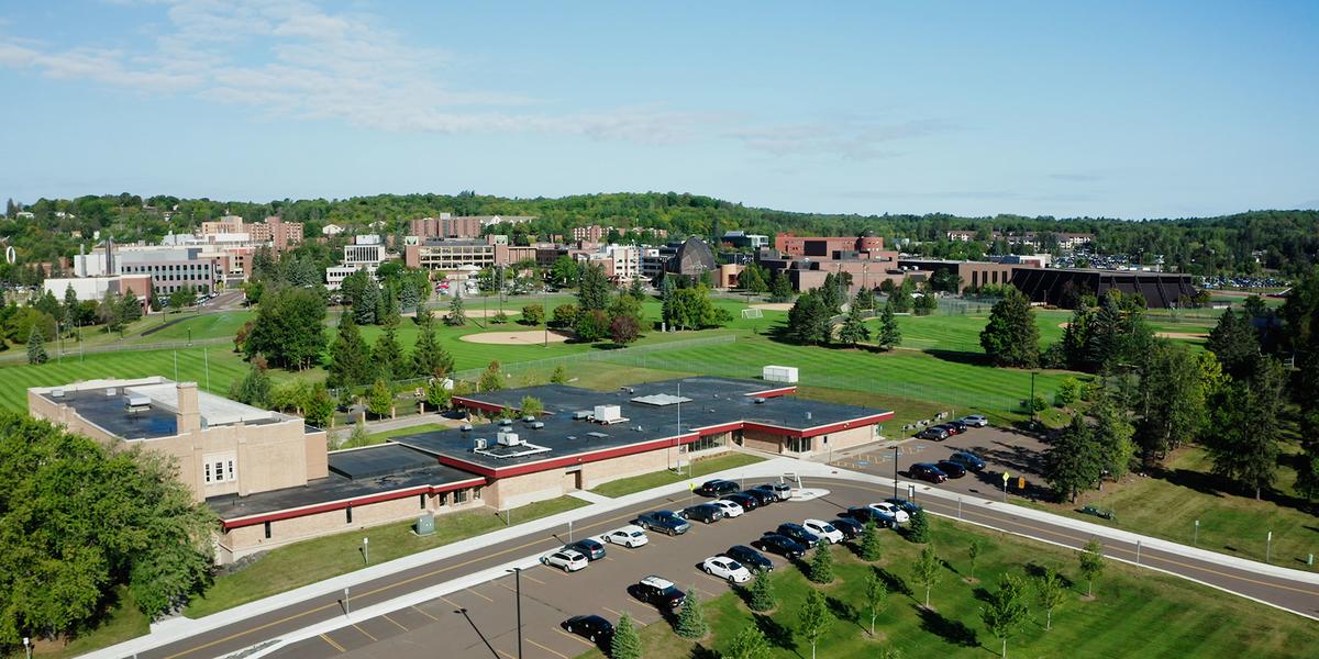 An aerial view of the Chester Park Building on the UMD campus. Green lawns and full trees with a parking lot with cars in front of the building and the rest of UMD campus in the background under a blue sky.