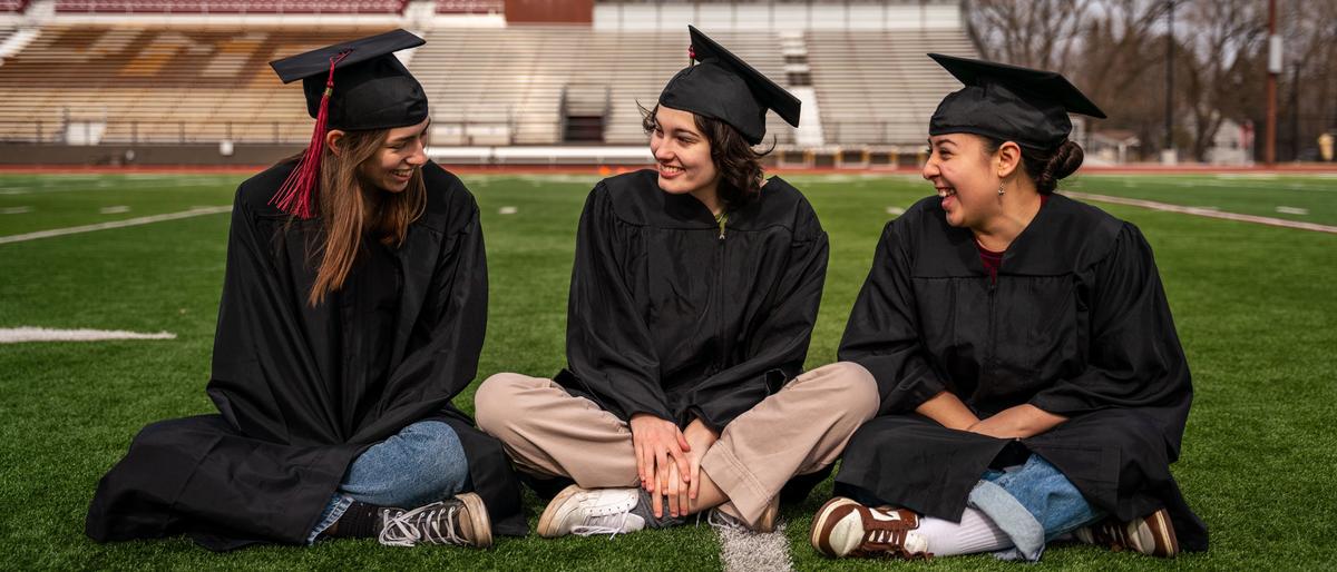 Three UMD students wearing graduation caps and gowns sit smiling and laughing on Griggs Field with bleachers in the background.