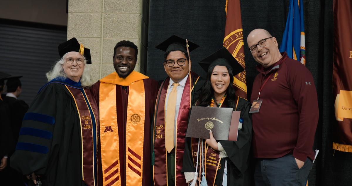 A group photo of people in graduation regalia in front of University of Minnesota flags.