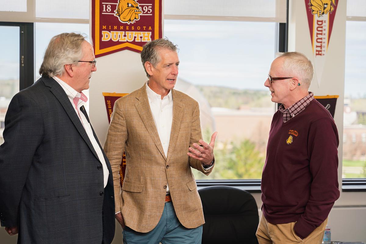 Three Chancellors standing discussing in a room with UMD pennants in front of a window overlooking the UMD campus.