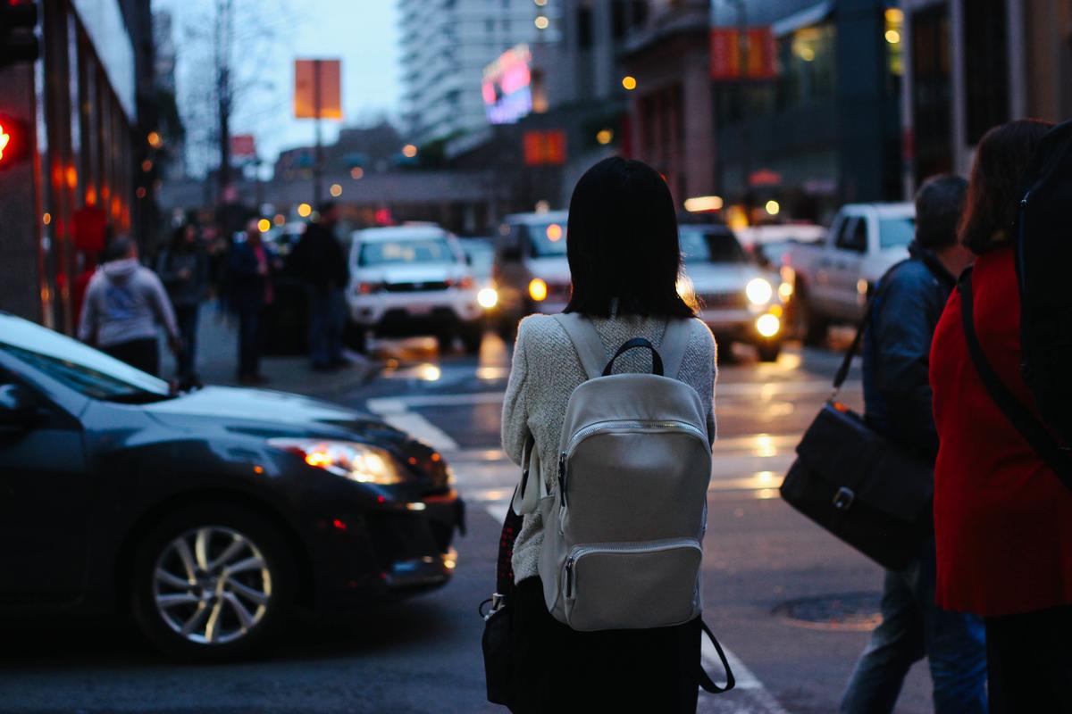 Young person seen from behind waiting to cross a busy street at twilight