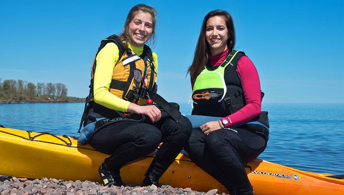 UMD alumna who paddled around Lake Superior sit by their kayaks on the shoreline