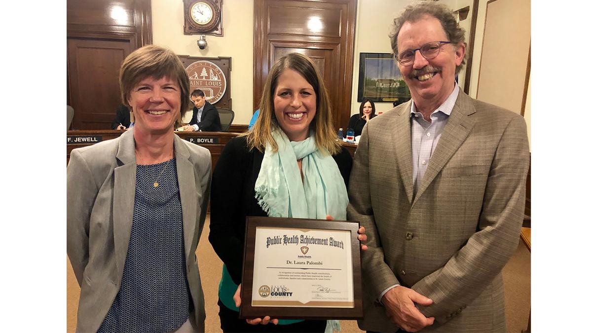 Dr. Laura Palombi (center) standing holding framed award, flanked by Amy Westbrook, director of Public Health on Laura's right, and Commissioner Frank Jewell, who chairs the Health and Human Service Committee.
