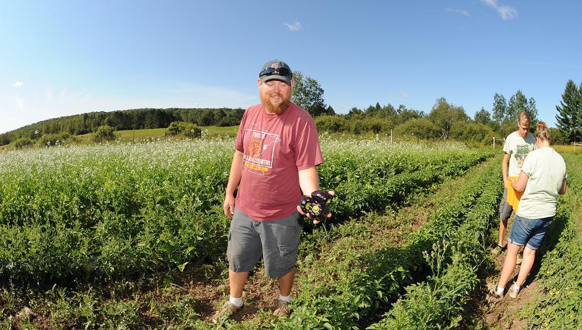 UMD's farm manager holds up a vegetable while students work on the farm in the background.