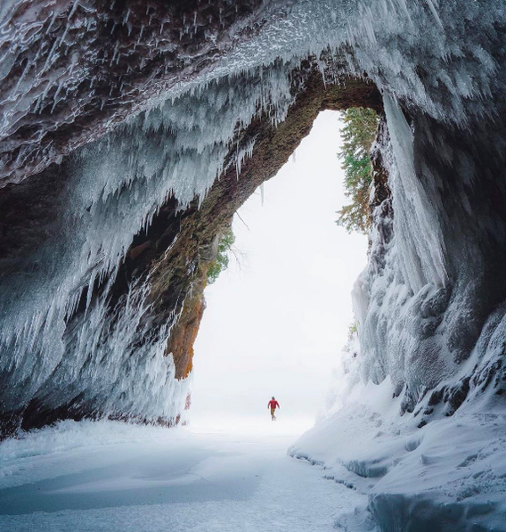 A tiny person wearing a red coat stands at the entrance of a very tall ice cave