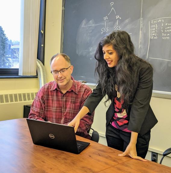 A UMD student with long black hair and a black blazer leans over a laptop while Ted Pedersen, PhD, looks on. There's a blackboard with equations in the background.