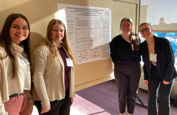 4 PETE students smiling in front of their research board titled " oxigen consumptions and substate use during sub-maximal exercise in females during the Follicular and luteal phases"