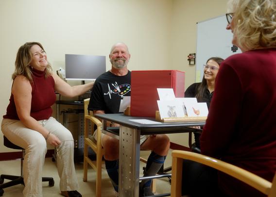 A therapy session with all four people smiling. Associate Professor Sharyl Samargia-Grivette has her hand on research participant Bill Mayo's back as Madelyn and Lynette look on.