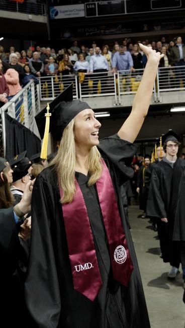 Amanda Fowler walking into the commencement ceremony, wearing a cap and gown, and smiling and waving at friends and family