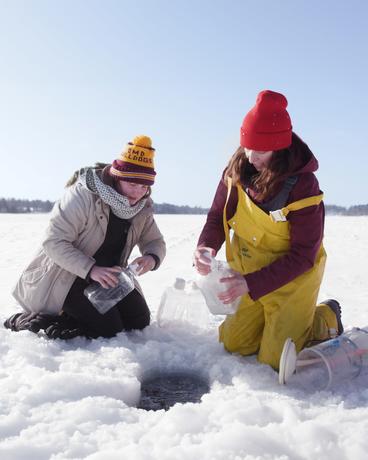 Two UMD students handle water sampling equipment over a hole in the ice on a frozen lake.