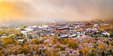 An aerial photo of campus with brightly lit clouds, fall color on the trees, and the light dusting of the first snow